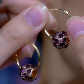 Pair of gold hoop earrings with purple crystal pavé ball charms held between fingers