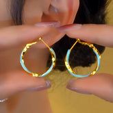 Gold and light blue twisted hoop earrings held near a person's ear