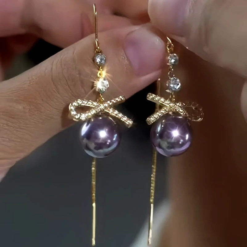 Pair of gold bow purple pearl crystal dangle drop earrings held in hand