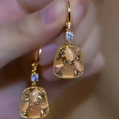 Hand holding pair of gold square cat eye dangle earrings with cubic zirconia accent