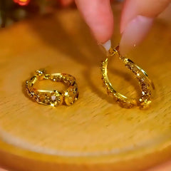 Sparkling twisted gold hoop earrings, one held by fingers on warm wooden surface