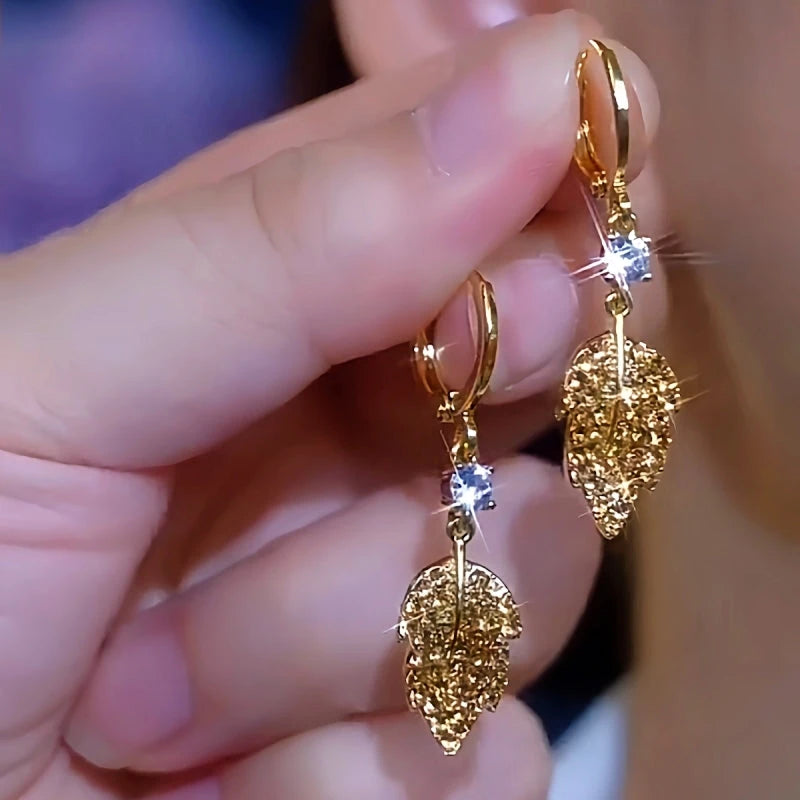 Pair of gold leaf dangle hoop earrings with clear crystal, held in hand