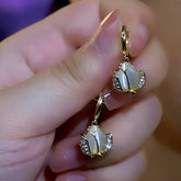 Hand holding pair of gold tulip dangle hoop earrings with white cat eye and crystal accents