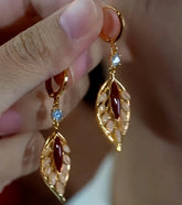 Hand holding pair of gold leaf dangle earrings with red agate and light blue gem accents