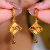 Gold rhombus drop earrings with orange cat's eye stones, held by manicured fingers