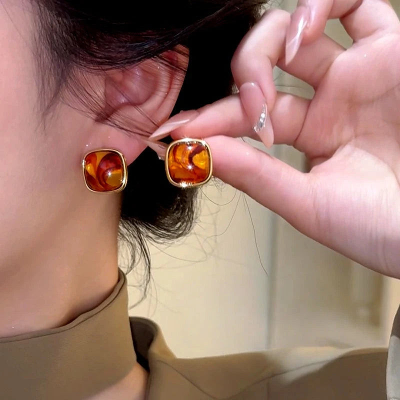 Amber swirl square gold stud earrings: one worn on ear, one held for size reference