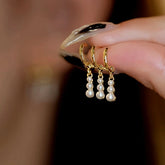 Three gold pearl drop huggie hoop earrings held by manicured hand