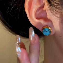 Round gold turquoise stud earring worn on an earlobe, manicured hand beside it