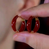 Pair of glossy deep red enamel gold-trimmed huggie hoop earrings held between fingers