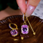 Purple emerald-cut crystal gold drop hoop earrings, one held over wooden dish