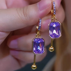 Pair of purple emerald-cut crystal gold drop hoop earrings held in hand