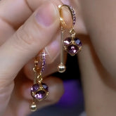 Pair of gold purple cubic crystal dangle huggie hoop earrings held in hand
