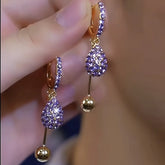 Pair of purple rhinestone teardrop gold ball dangle hoop earrings held in hand