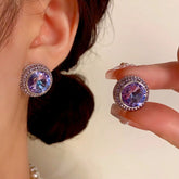 Round purple-blue crystal halo stud earrings, one worn on ear one held by hand