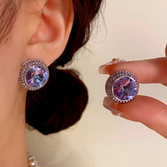 Round purple-blue crystal halo stud earrings, one worn on ear one held by hand