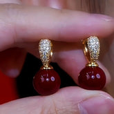 Pair of gold crystal pave huggie hoop earrings with red bead drop held between fingers