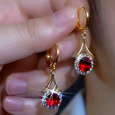Pair of gold huggie drop earrings with ruby red gem and crystal halo, held in hand