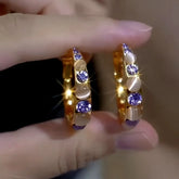 Pair of gold half hoop earrings with purple crystals and opal stones held by fingers