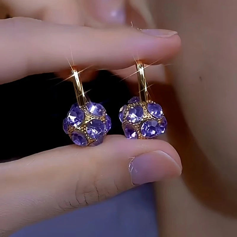 Pair of purple crystal ball gold hoop earrings held between fingers