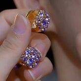 Pair of gold purple rhinestone cluster huggie earrings held in hand