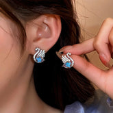 Sparkly rhinestone swan stud earrings with blue accent, one worn on ear one held