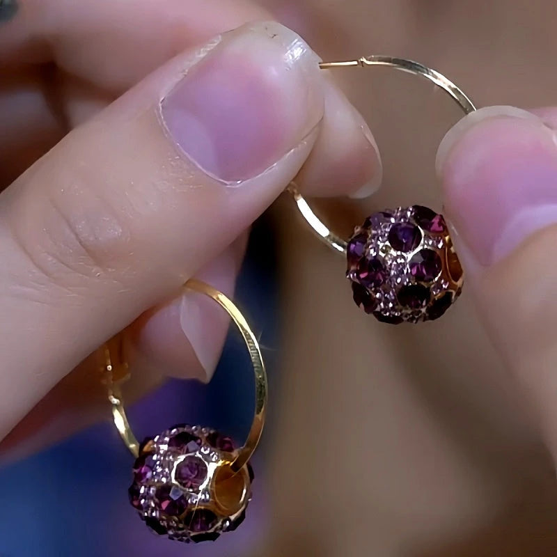 Pair of gold hoop earrings with purple crystal pavé ball charms held between fingers