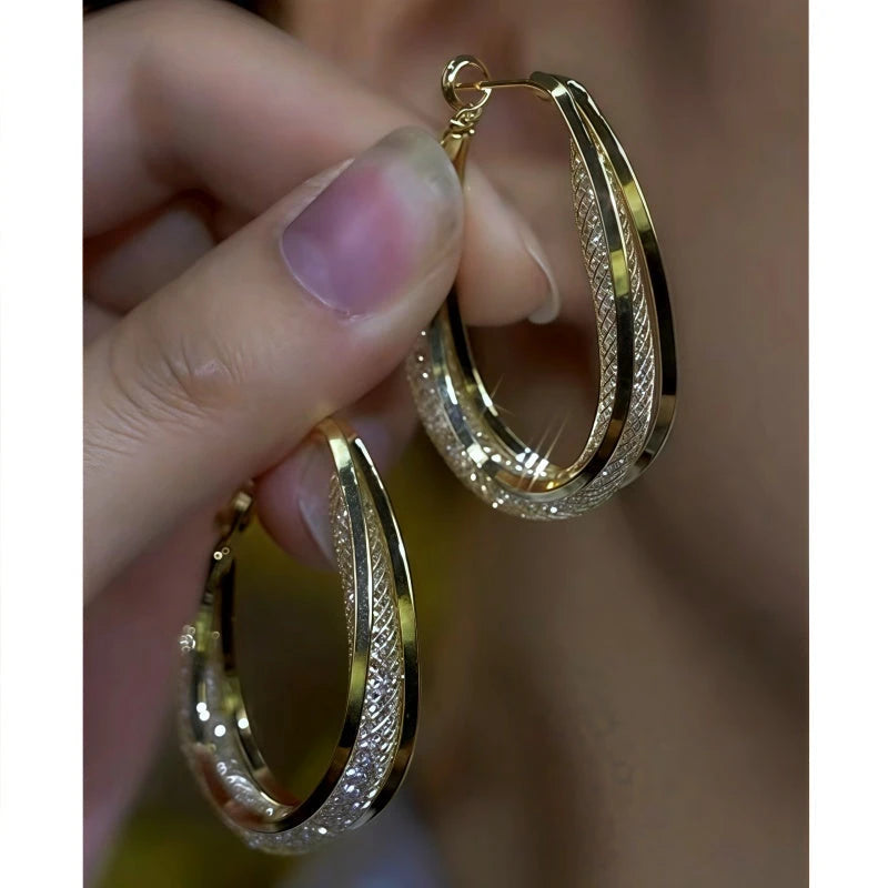 Hand holding pair of gold textured oval hoop earrings with shimmer detailing