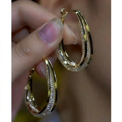 Hand holding pair of gold textured oval hoop earrings with shimmer detailing