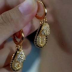 Hand holding pair of gold two-tier textured teardrop hoop dangle earrings with black trim