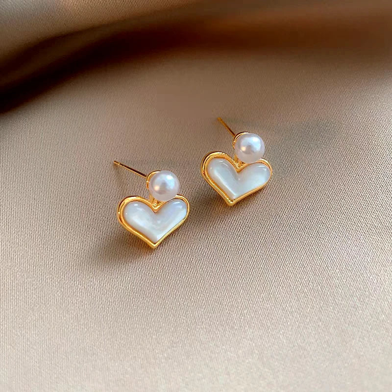 Pair of gold mother of pearl heart stud earrings with pearl accent on beige satin fabric
