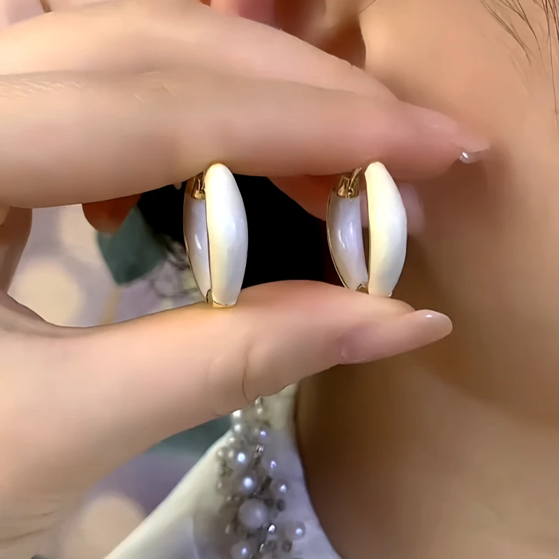 Pair of white enamel gold-tone huggie hoop earrings held in hand