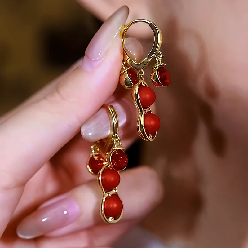 Pair of gold red peanut bead dangle hoop earrings held by manicured hand