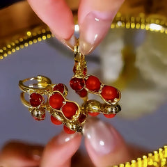 Gold peanut-shaped hoop earrings with red gemstones, held by fingers on reflective surface