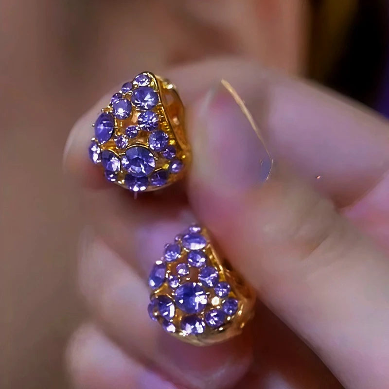 Pair of gold-tone purple rhinestone triangular stud earrings held in hand