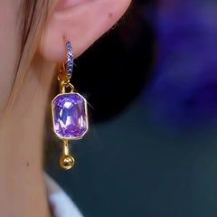 Gold dangle earring with purple pave CZ hoop, cushion-cut purple gem, and gold ball, worn on ear
