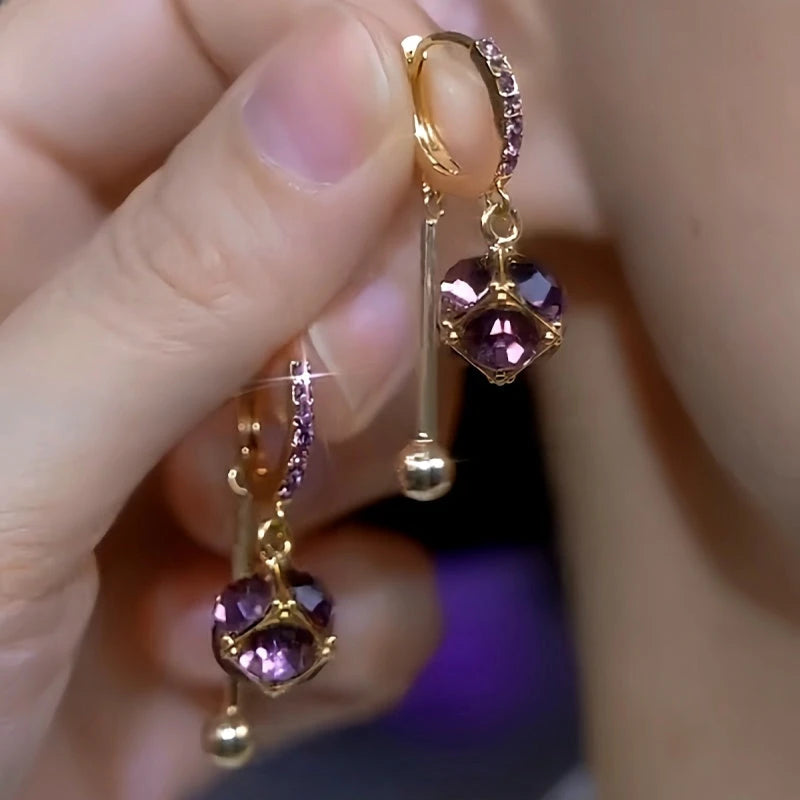 Pair of gold purple cubic crystal dangle huggie hoop earrings held in hand
