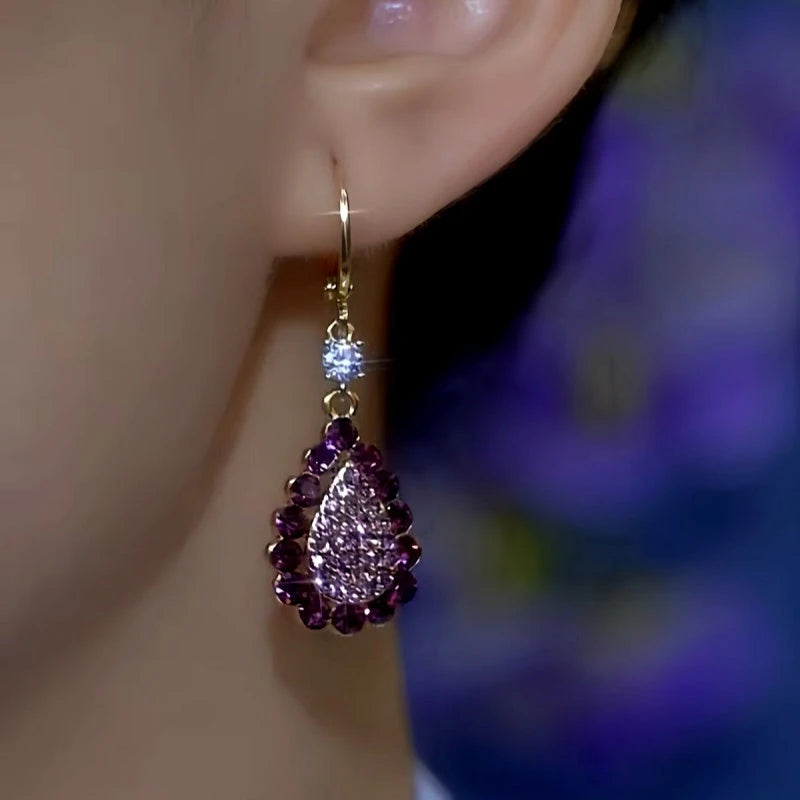 Close-up of a purple teardrop rhinestone dangle earring with gold hoop on woman's ear