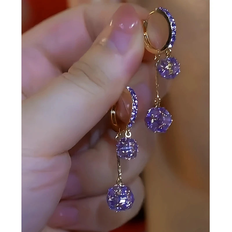 Hand holding pair of gold-tone purple crystal dangling hoop earrings
