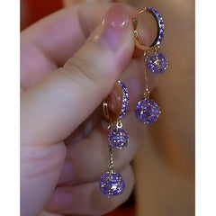 Hand holding pair of gold-tone purple crystal dangling hoop earrings