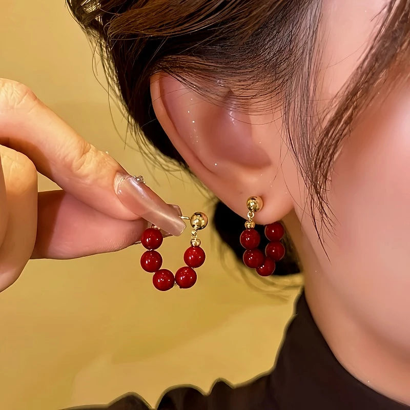 Women's red beaded gold stud C-shaped hoop earrings, hand holding one beside worn earring