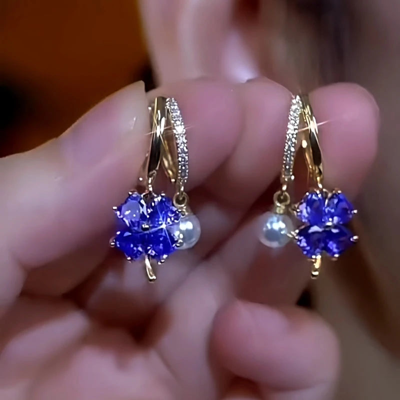 Pair of blue four-leaf clover CZ pearl pave double gold huggie hoop earrings, held in fingers