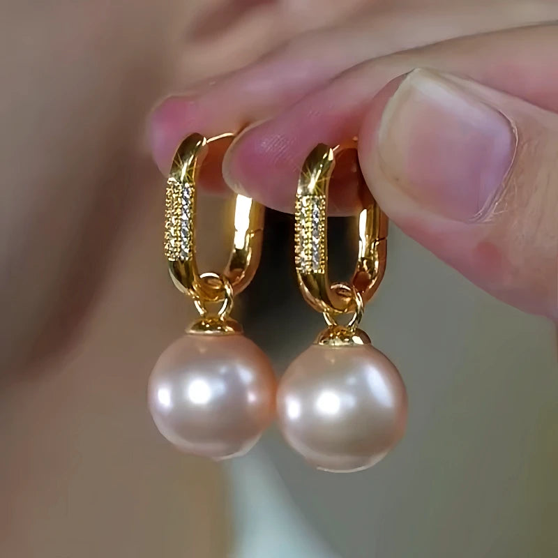 Pair of gold pave pearl drop huggie hoop earrings held between fingers