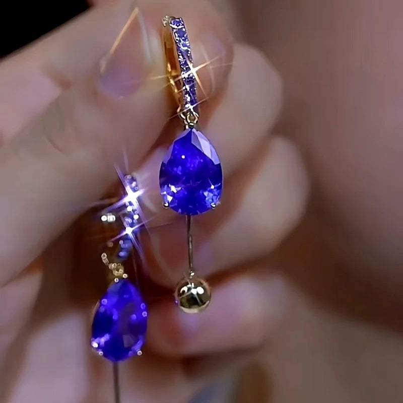 Pair of purple teardrop crystal gold ball dangle hoop earrings held in hand