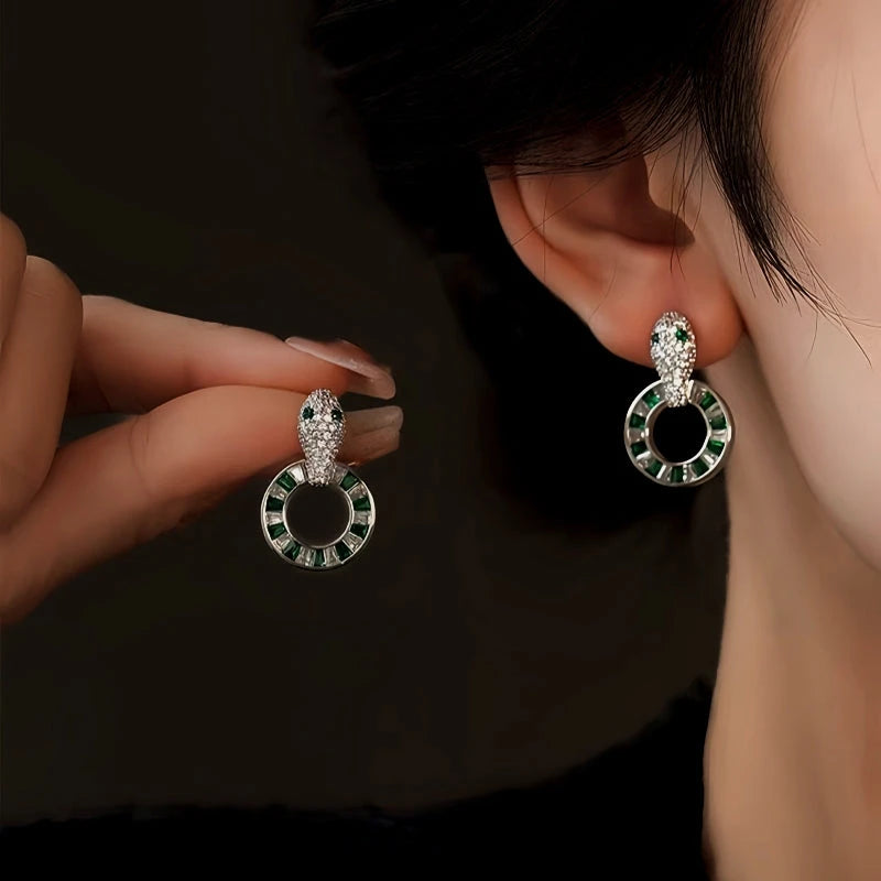 Silver snake head drop earrings with green gem circle, worn on ear and held in hand