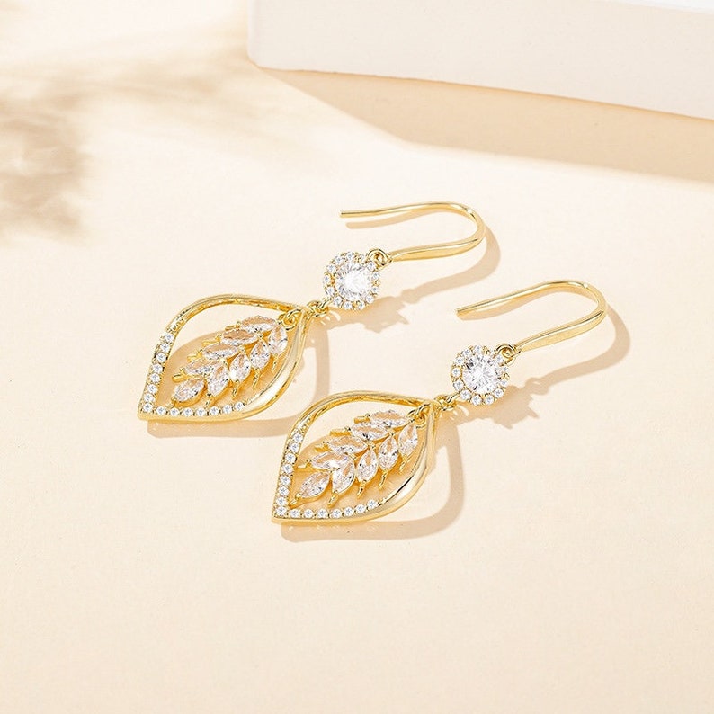 Pair of gold leaf design CZ drop earrings laid flat on warm beige surface
