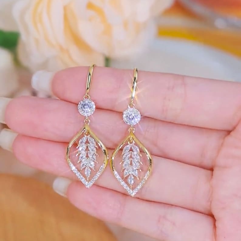 Gold CZ leaf drop earrings held on a hand, lifestyle product shot